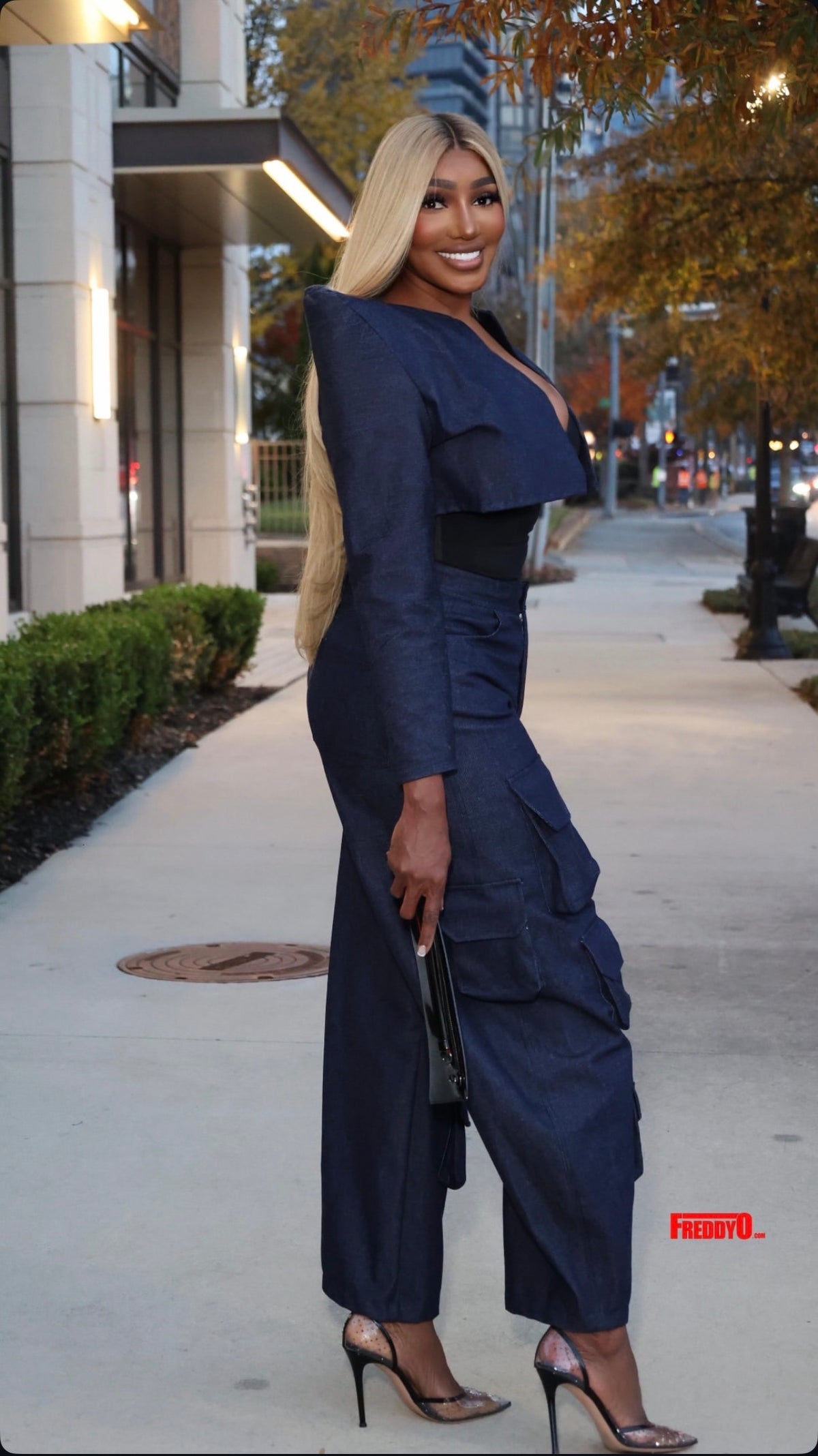 A woman posing on an outdoor city sidewalk wearing a navy blue ruffled jumpsuit with wide-leg pants, paired with clear high-heeled sandals and holding a black clutch.